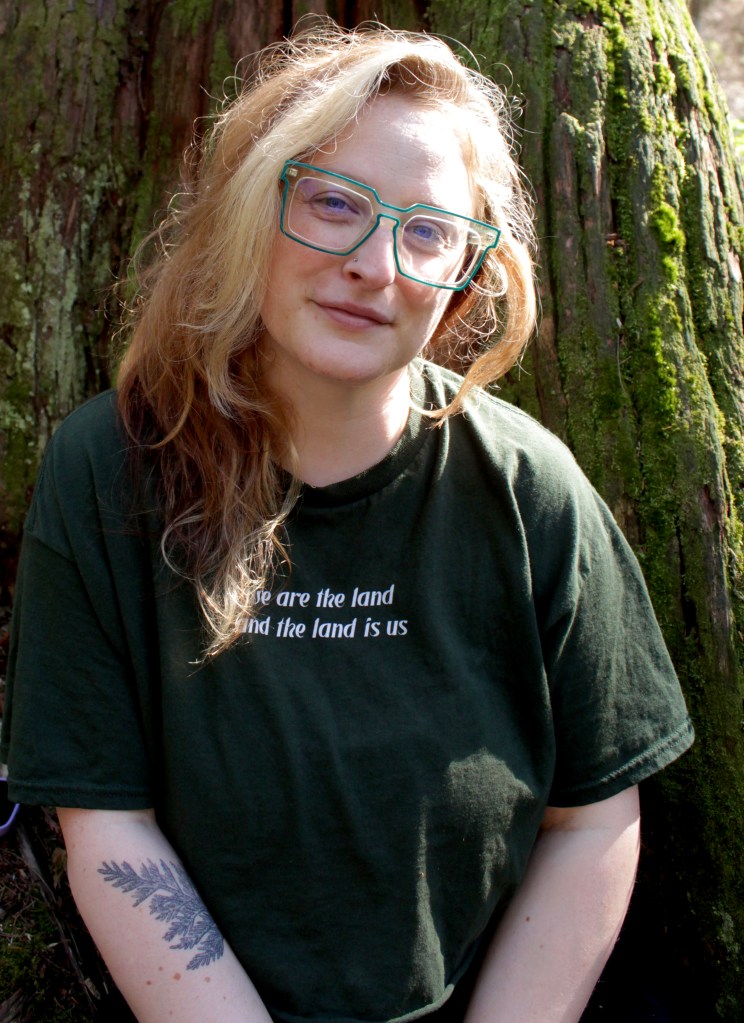A photography of Laken sitting against an old growth cedar, covered in lichen and moss. Laken has chest length hair, with blonde stripes in the front, copper red colour, and chocolate streaks underneath. They are wearing blue lined square shaped glasses and a dark forest green shirt that reads "we are the land and the land is us". On Laken's left arm is a tattoo of a cedar branch.